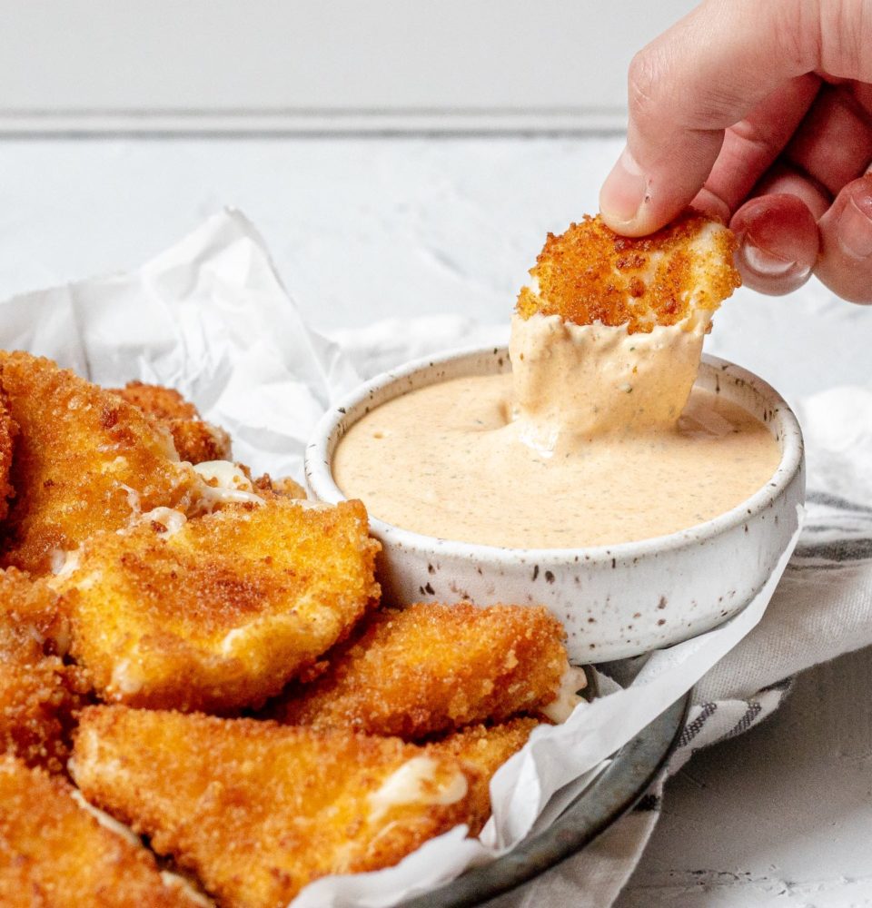 Breaded and fried cheese being dipped into a white ceramic bowl filled with a creamy dip.