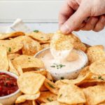 A person dipping a seasoned chip into a bowl of dip, made with Oaxaca cheese, that is surrounded by chips.