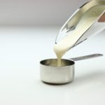 A silver scoop pouring concentrated flavoring paste into a silver measuring cup.