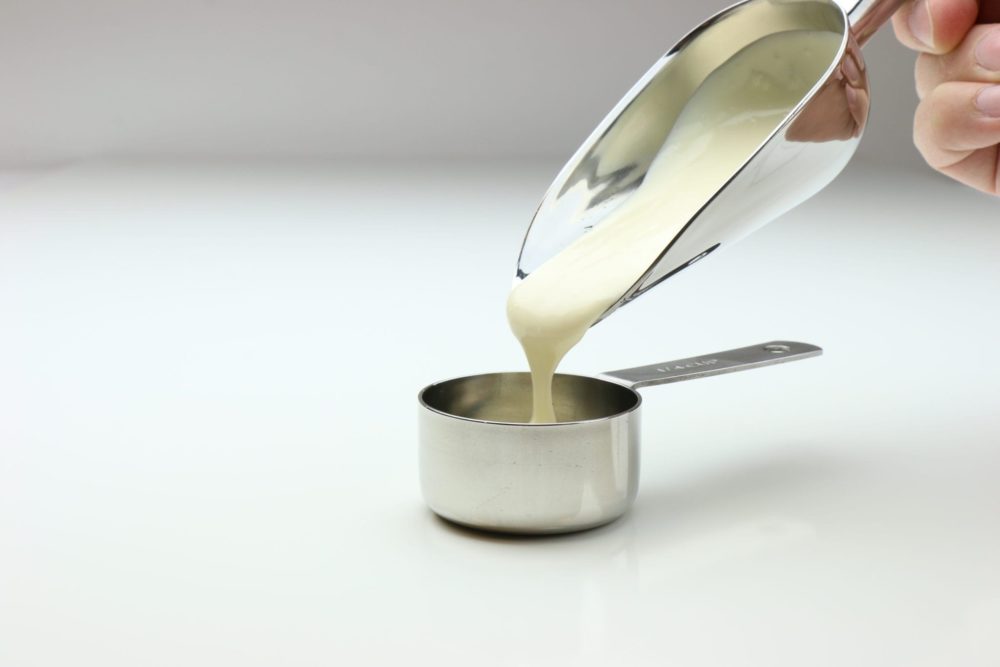 A silver scoop pouring concentrated flavoring paste into a silver measuring cup.