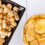 Popcorn sitting on a rectangular plate next to two bowls of seasoned potato chips.