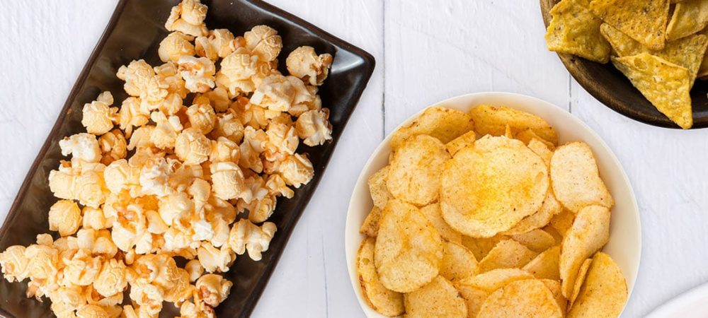 Popcorn sitting on a rectangular plate next to two bowls of seasoned potato chips.