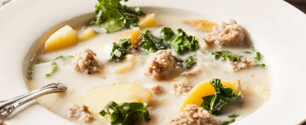 A bowl of spicy sausage and potato soup with a silver spoon sitting inside of it.